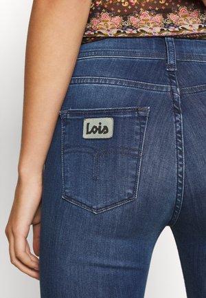 Ciemnoniebieskie dżinsy z denimu mają tylną kieszeń z szarą etykietą, na której czarnym pismem zapisano "Lois". Tkanina jest gładka z delikatnym połyskiem.