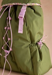 Olivovo zelený batoh s ružovými kovaniami, vybavený kvetinovým lemom, dizajnom s falošným šnurovaním a bočným elastickým vreckom na uloženie vecí.