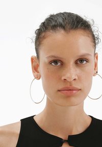 Joven mujer con pelo rizado y oscuro recogido, lleva pendientes de aro grandes y una camiseta negra con un pequeño cierre frontal.