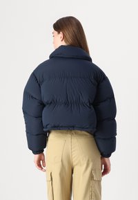 Navyblauwe gewatteerde jas met een hoge kraag, dikke quilted secties en elastische manchetten. Draagend over kaki broeken, bekeken van achteren.