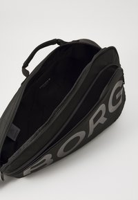 Mochila preta feita de tecido durável, com um grande bolso frontal que exibe um ousado logo cinza. O compartimento com fecho revela espaço de armazenamento adicional.