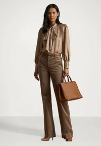 Ženska oblečena v rjavo vzorčasto bluzo s kravato, rjave hlače visoke pasu, rjave petke in drži rjav usnjen ročno torbico.