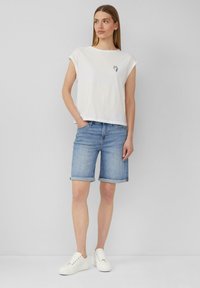 Weißes Baumwoll-T-Shirt mit hochgerollten Ärmeln und einem kleinen Grafikdesign, kombiniert mit hellblauen Jeansshorts und weißen Sneakern. Einfaches, lässiges Outfit.