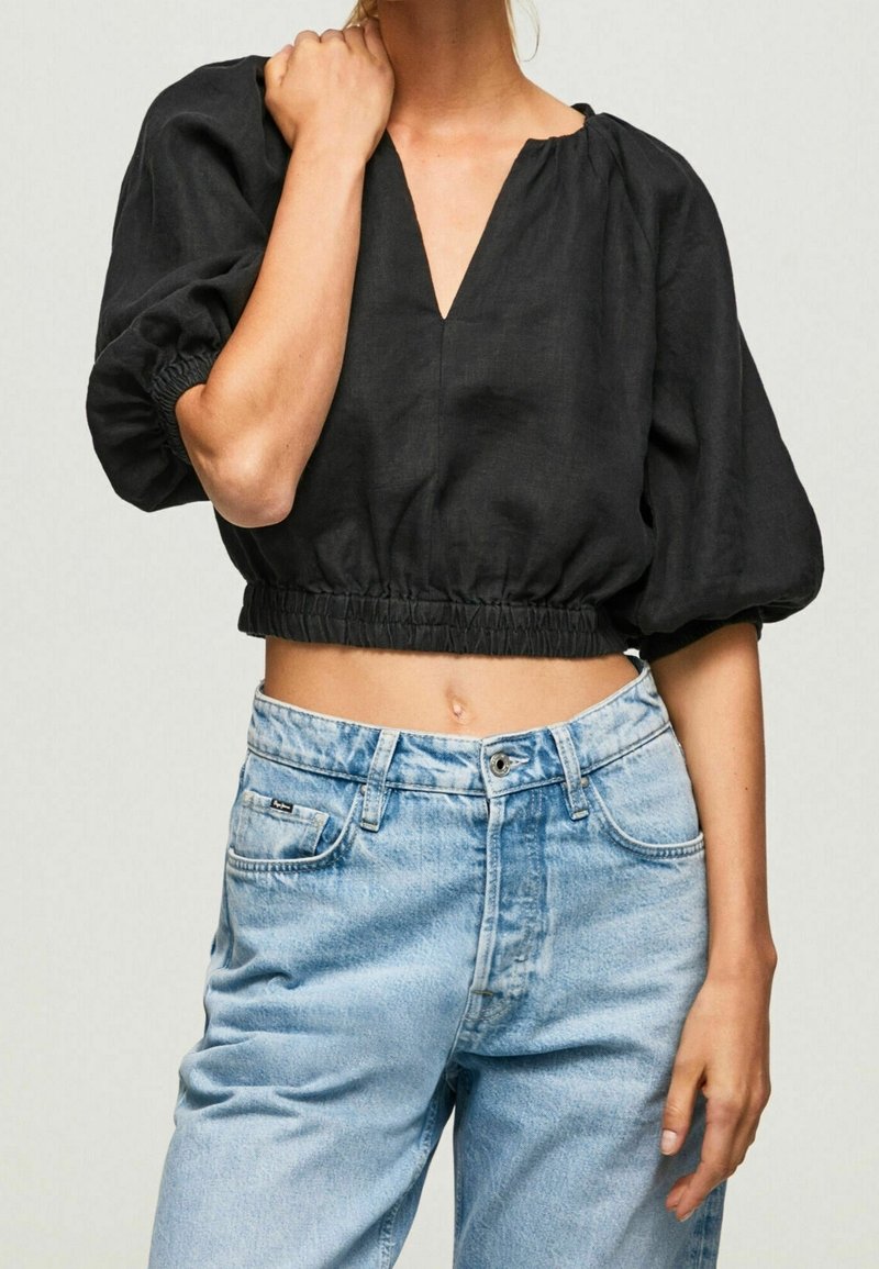 Femme portant un crop top noir à manches bouffantes et col en V, associé à un jean bleu clair taille haute sur fond uni.