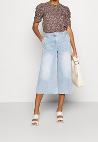 Femme portant un blouse à manches bouffantes florale rentrée dans un jean cropped large bleu clair, tenant un sac à main beige et des sandales à talons blanches.