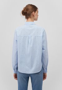 Himmelblaues Hemd mit Knopfleiste aus leichtem Stoff, mit kleinen blauen Punkten und einem Rückeneinsatz mit einer Mittelnaht. Lange Ärmel mit Manschetten.