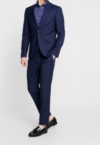 Granatowy garnitur w wzory z dopasowanym marynarką i zwężanymi spodniami, połączony z kwiecistą koszulą na guziki i czarnymi loafersami. Gładka faktura tkaniny.