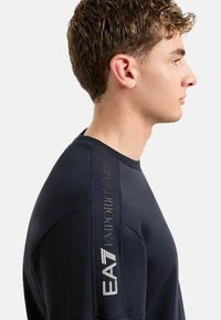 Giovane in profilo che indossa un felpa nera EA7 Emporio Armani con il logo stampato sulla manica superiore.