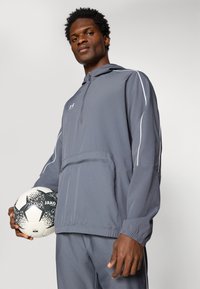 Siva lagana jakna s kapuljačom s reflektirajućim detaljima, s polu-zip zatvaranjem sprijeda i kenguru džepom. Model drži crno-bijelu nogometnu loptu.