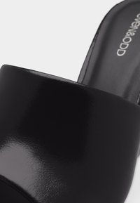 Gros plan d'une mule noire brillante à enfiler avec une large bande avant et la marque visible "Steven & ODD" sur la semelle intérieure.