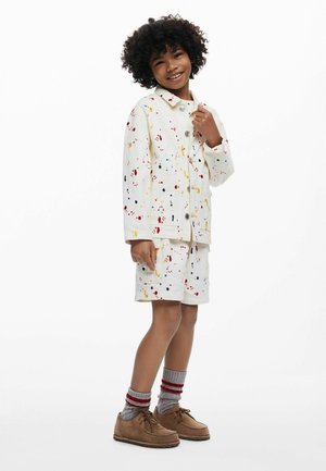 Balta džinsinė striukė ir šortai su spalvotų dažų purslų raštais. Rudi batai su tekstūruotomis padomis ir pilkos kojinės su raudonomis juostelėmis.