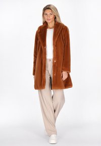 Kvinde der går, iført en brun pelsjakke over en hvid top, beige bukser og hvide sneakers, mod en ensfarvet hvid baggrund.