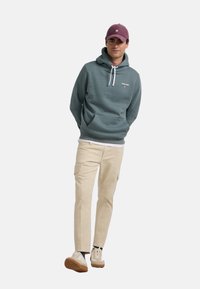 Sudadera verde con cordones blancos y logo, combinada con pantalones cargo beige. El personaje lleva zapatillas crema y una gorra morada.