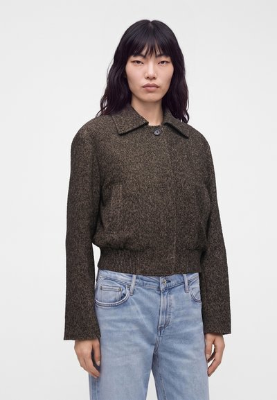 Femme aux longs cheveux noirs portant une veste texturée marron foncé et un jean bleu clair, debout devant un fond blanc uni.