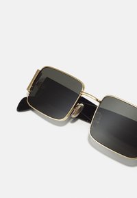 Lunettes de soleil carrées avec verres sombres, monture en métal doré et branches en plastique noir dotées de plaquettes de nez décoratives. Design épuré avec des accents élégants.