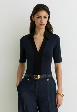 Žena oblečená v tmavomodrom topu s čiernym golierom a hlbokým výstrihom do V, v kombinácii s vysokým pásom tmavých riflí, čiernym opaskom a zlatým náramkom s retiazkou.