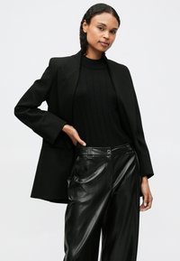 Blazer preto ajustado sobre um suéter preto canelado, combinado com calças de pele sintética pretas de cintura alta. Texturas suaves e design estruturado.