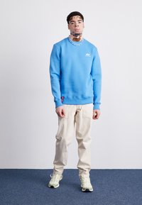 Blå sweatshirt med ribbede manchetter og kant, crema cargo bukser og olivengrønne sneakers. Har et logo på brystet og en sidetags.