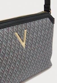 Musta ja harmaa geometrinen kuvioitu laukku, jossa on sileä pinta, kullanvärinen "V"-koriste ja vetoketjullinen suljin.