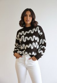 Kobieta o ciemnych włosach do ramion nosi ciemny sweter z białymi zygzakowymi wzorami oraz białe spodnie z wysokim stanem, pozując z dłońmi w kieszeniach.