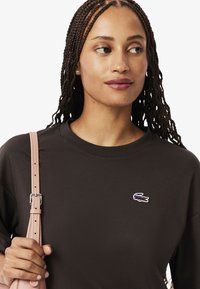 T-shirt in cotone marrone con collo a giro e maniche corte, caratterizzato da un logo ricamato sul petto e una borsa a tracolla rosa.