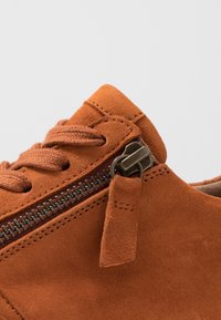 Bruna mockaskor med tjocka bruna snören och en metalldragkedja på sidan, med en mockadragslapp framför en enkel bakgrund.