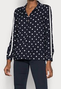 Blusa a pois blu navy con pois bianchi, chiusura con bottoni frontale, maniche lunghe e strisce laterali bianche, realizzata in un tessuto leggero.