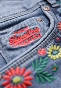 Džínovina s modrým odstínem, zdobená červenou a barevnou květinovou výšivkou, s výrazně vyšitou červenou logem „Superdry“.