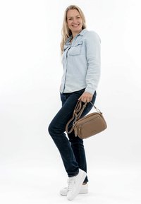 Borsa a tracolla marrone con finitura testurizzata, tasca con zip e tracolla regolabile. Indossata con una camicia di denim chiara, jeans scuri e sneakers bianche.