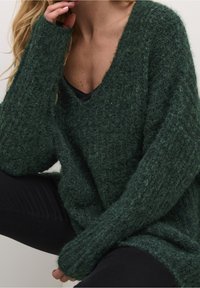Oversized grön stickad tröja med V-ringning, ribbad textur och långa ärmar. Buren över en svart topp, mot en neutral bakgrund.