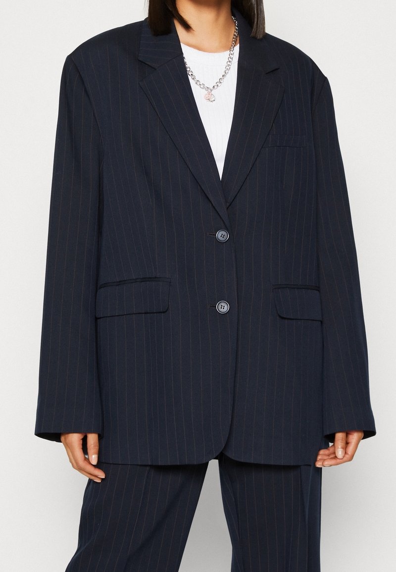 Blazer blu navy con righe sottili e vestibilità rilassata, con revers a punta, due bottoni e tasche laterali. Indossato sopra una maglietta bianca.