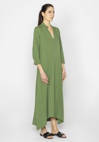 Długa zielona sukienka z dekoltem w serek i rękawami o długości trzech czwartych, charakteryzująca się swobodnym krojem i asymetrycznym dołem. Noszona z czarnymi klapkami.