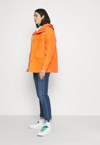 Pessoa vestindo um casaco com capuz laranja brilhante, jeans azuis com as mangas arregaçadas, tênis brancos com cadarços verdes, de pé de lado contra um fundo branco.