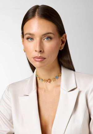 Kobieta o prostych, ciemnych włosach, ubrana w biały żakiet oraz kolorowy naszyjnik w kwiatowy wzór i pasujące kolczyki, patrząca prosto przed siebie z neutralnym wyrazem twarzy.