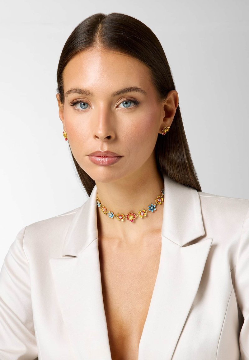 Femme aux cheveux raides et foncés, portant un blazer blanc, un collier floral coloré et des boucles d'oreilles assorties, regardant droit devant avec une expression neutre.
