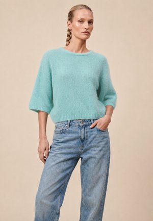 Femme aux cheveux tressés portant un pull duveteux bleu clair à manches trois-quarts et un jean bleu délavé ample sur un fond beige.