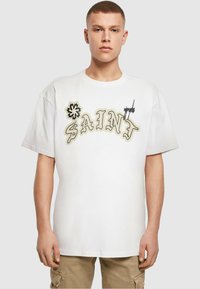 Balta medvilninė marškinė su ryškiu smėlio spalvos „SAINT“ tekstu ir gėlių grafikomis, laisvu siluetu ir apvaliu iškirpimu.