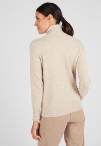 Beige coltrui met een geribde kraag en manchetten, gemaakt van een zachte, gestructureerde stof. De achterkant heeft een rechte zoom.