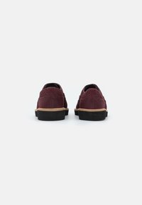 Une paire de chaussures brogues en cuir bordeaux avec des semelles noires et des semelles intermédiaires brunes, vues de l'arrière sur un fond blanc.