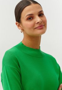Groene trui met hoge hals, zachte textuur en ribbels. Model draagt gouden oorringen. Minimalistische achtergrond.