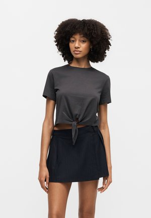 Mladá žena s kučeravými vlasmi, ktorá má na sebe krátky čierny viazaný crop top a čiernu mini sukňu na jednoduchom pozadí.