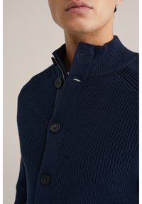 Mörkblå stickad tröja med hög krage, utrustad med dragkedja framtill och fyra svarta knappar. Ribbstickad textur med kontrasterande sömnadsdetaljer.