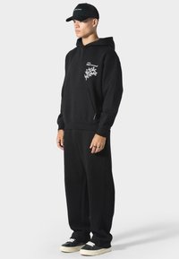 Zwarte hoodie met een voorzak en witte grafische prints, gecombineerd met bijpassende zwarte trainingsbroek en zwarte sneakers met witte zolen.