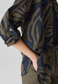 Maglietta blu navy con motivi astratti di foglie verdi, con maniche arrotolate e dettaglio con bottoni. Fit morbido, abbinata a pantaloni di colore verde oliva chiaro.