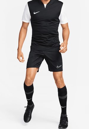 Homme portant un uniforme de football Nike noir et blanc, short noir, chaussettes longues noires et crampons noirs en position active.