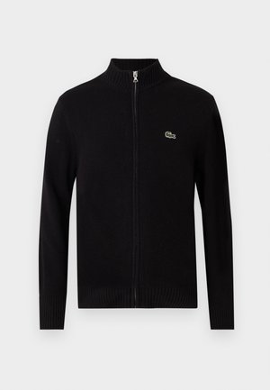 Zwarte ritssluiting cardigan van ribgebreid materiaal, met een hoge kraag en een klein groen logo op de linkerborst.