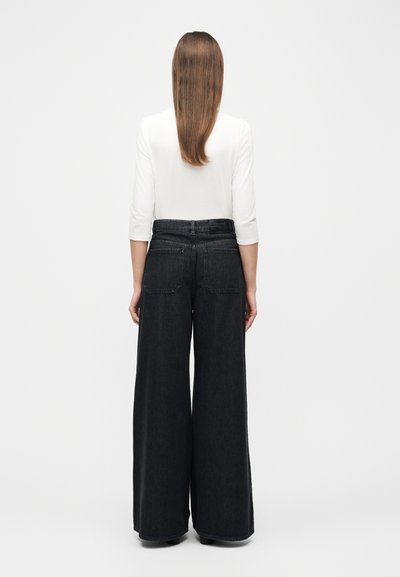 Γυναίκα με μακριά ίσια μαλλιά φοράει λευκή μπλούζα με μανίκια τριών τετάρτων και φαρδιά μαύρα τζιν ψηλόμεσα παντελόνια, γυρισμένη πλάτη σε λευκό φόντο.