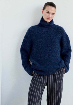 Mujer que lleva un suéter de cuello alto azul marino y peludo y pantalones negros con rayas verticales blancas, de pie con las manos en los bolsillos.