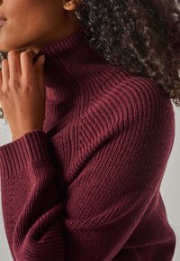 Frau mit dunklen lockigen Haaren, die einen kastanienbraunen, gerippten Rollkragenpullover trägt und mit der linken Hand ihr Kinn berührt.