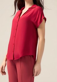 Blouse rouge à manches courtes avec un devant boutonné, un col et une texture lisse. L'ourlet est légèrement plus long à l'arrière, complétant un pantalon assorti.